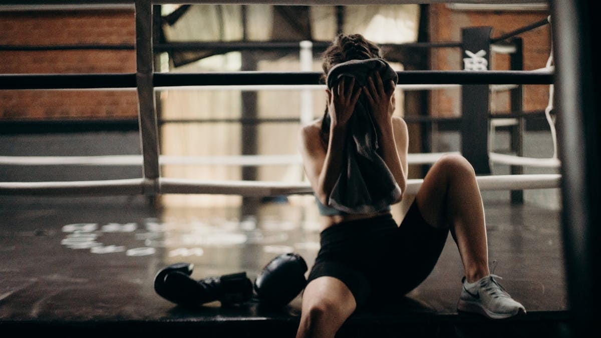 A female boxer sitting in a gym ring, resting and wiping sweat with a towel after a workout.